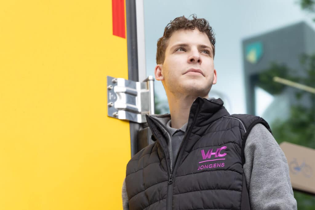 Nieuwe bedrijfskleding VHC Jongens met trots gedragen door bezorger in body warmer naast vrachtwagen