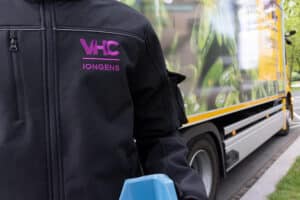 Nieuwe bedrijfskleding VHC Jongens als zwart vest gedragen door bezorger met vrachtwagen op de achtergrond